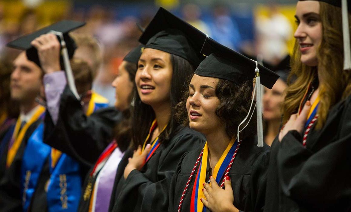 Students in graduation caps and gowns stand with hands over hearts