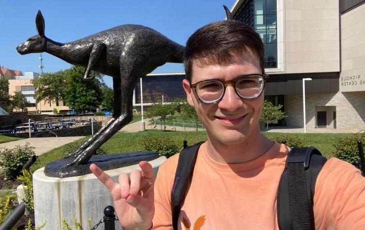 Brett Boyer takes a selfie outside on campus, in front of the Roo Statue and Miller Nichols Library. He smiles and holds up a hand in a Roo Up gesture.