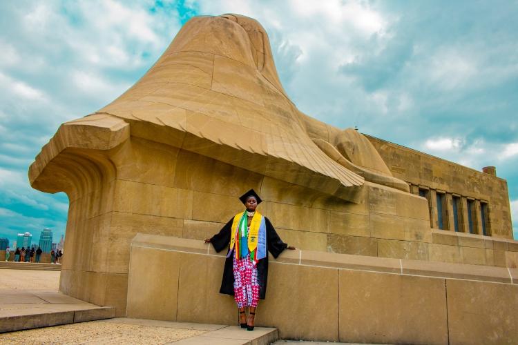 护理系毕业生可可Ndipagbor穿着毕业礼服站在自由纪念碑的亚述狮身人面像前