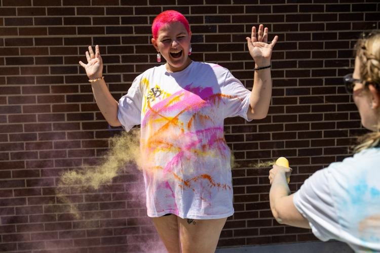 A student stands with arms out as people toss color powder on their shirt