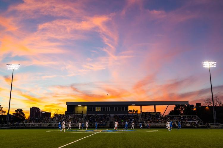 the men's soccer team plays rockhurst with the stands of Durwood in the background and a sunset