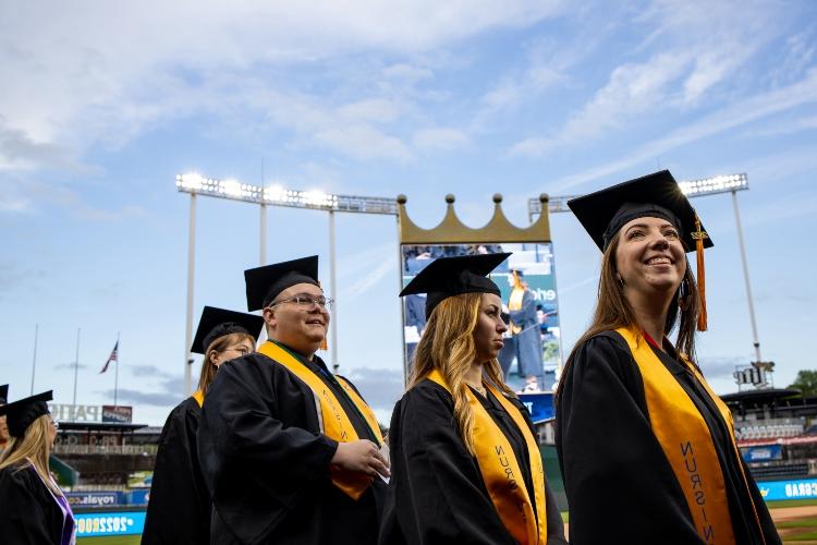 Three students stand in cap and gowns with Kauffman Stadium's Crown Vision screen in the background
