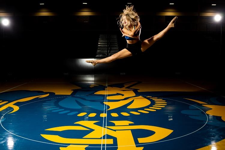 A dancer leaps in the air with her legs in a splits position. Below her is the Roo that is painted on the floor of the Swinney basketball court.