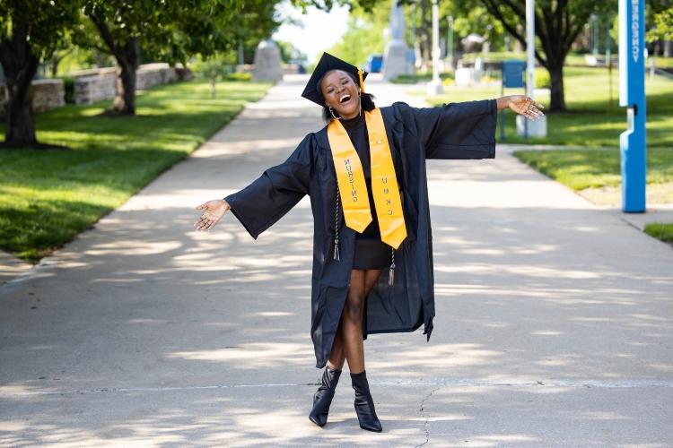 A graduate stands joyous on university walkway with one foot over the other, arms stretched out and head tilted, smiling