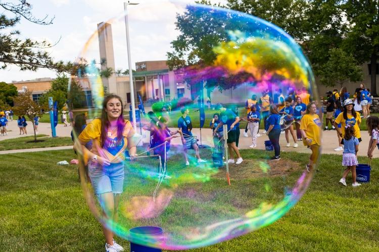 A translucent bubble is in the foreground with a student behind it, holding a bubble wand and smiling on a sunny university walkway