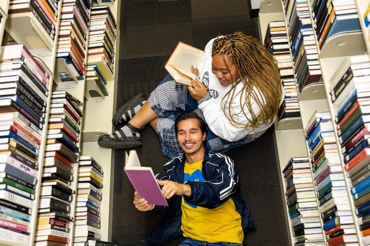 The camera angle is above two students sitting between two rows of books at the library. One is seated on the floor reading, while the other is laying with his head on their knee reading.