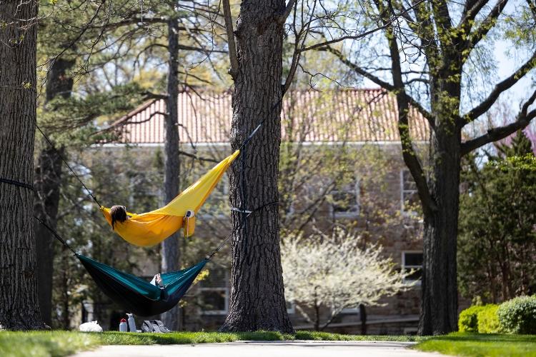 Two students lay in hammocks that are hung between trees on the quad, with one hammock hanging above the other