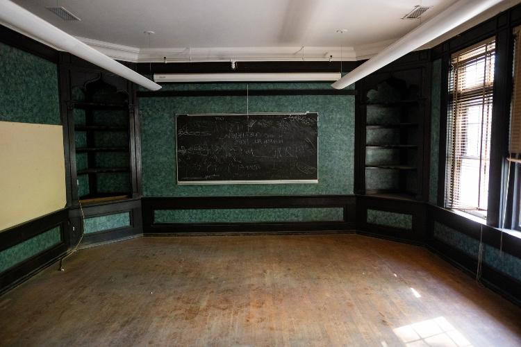 一个房间的广角镜头，后面墙上有一块黑色的黑板. 角落里有黑色的书架. 墙上装饰着华丽的绿色墙纸
