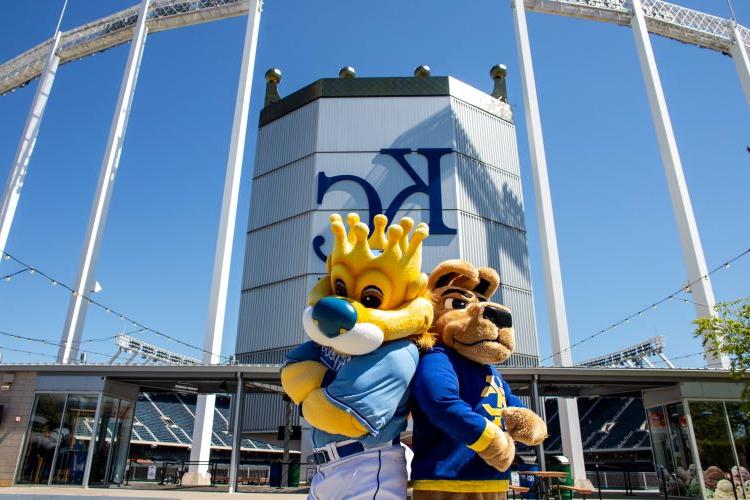 KC Roo and Sluggerrr pose standing back-to-back in front of the CrownVision board at Kauffman Stadium