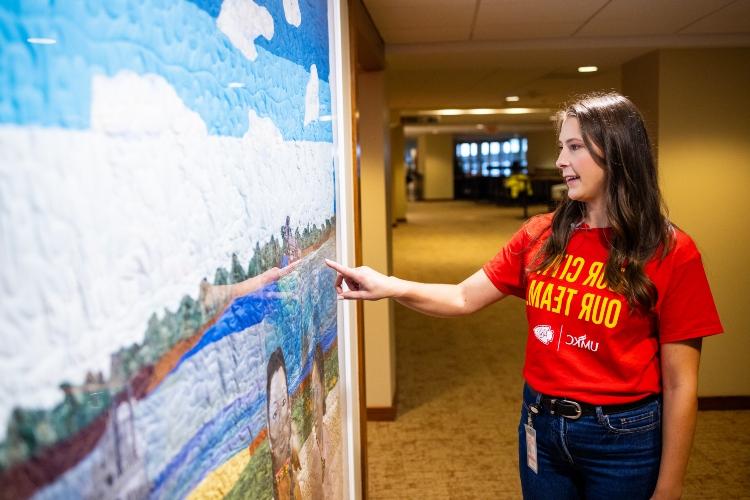 Meghan Jaben points to a quilt hanging inside Arrowhead as part of the Arrowhead Art Collection while wearing a red UMKC and Chiefs shirt that says "Our City Our Team"