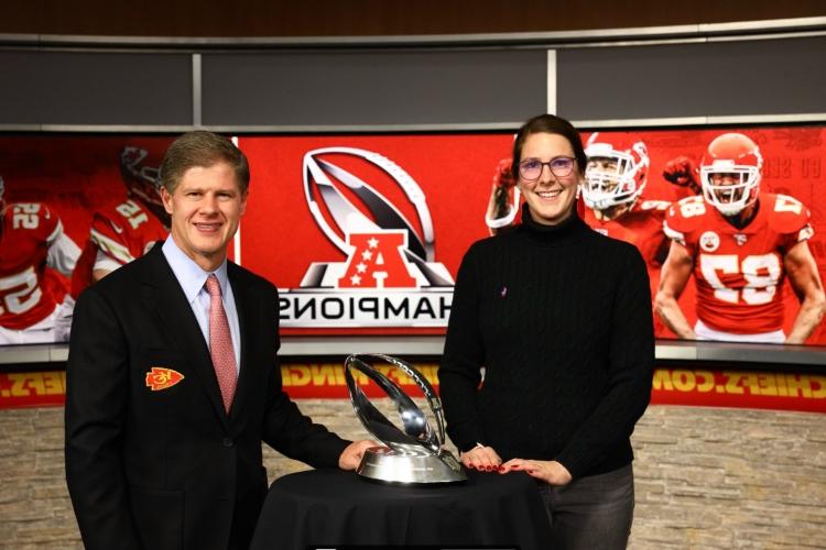 Meghan Jaben stands with the CEO of the Chiefs, Clark Hunt, with the AFC Championship Larmar Hunt trophy sitting on a table between them. Behind them a giant red Chiefs background has a photo of Travis Kelce yelling and an illustration of the Lamar Hunt trophy with the words, "AFC Champions"