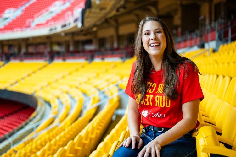 Meghan Jaben smiles seated in the yellow club seats at Arrowhead with a red shirt that says "Our City, Our Team" and the UMKC and Chiefs logos