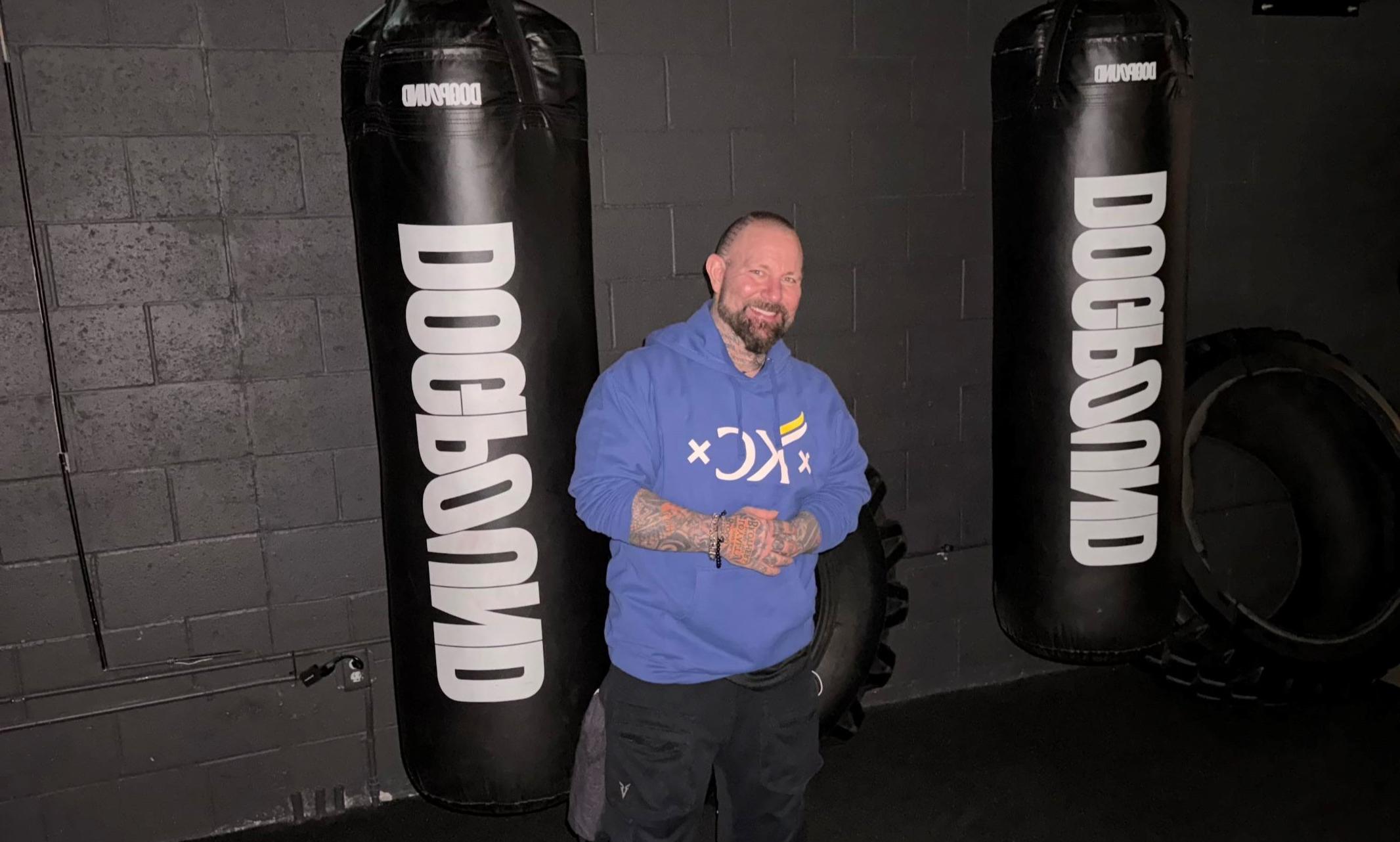 Taylor Swift and other celebrities' fitness trainer UMKC Alum Kirk Myers wearing a blue KC Mobb Made Sweatshirt in front of boxing sandbags at his gym dogpound