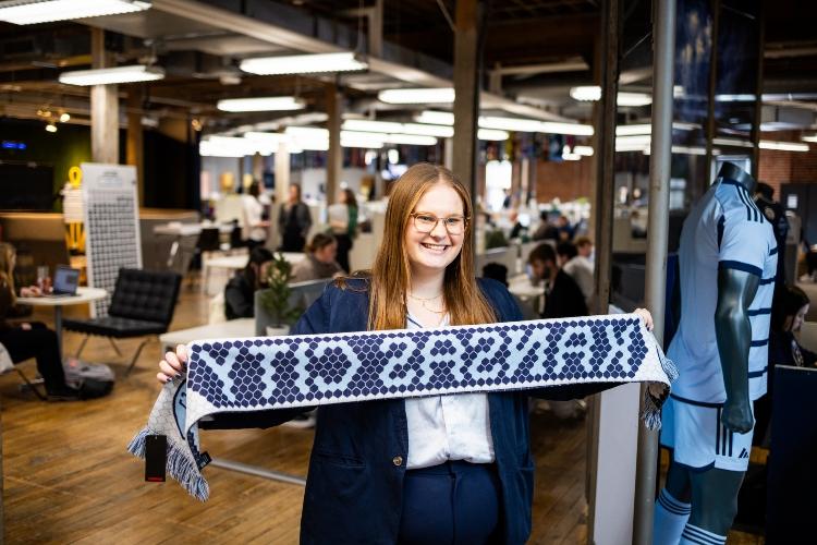 Annmarie Orlando stands smiling inside the Sporting KC offices holding a blue scarf that says "Kansas City" out in front of her