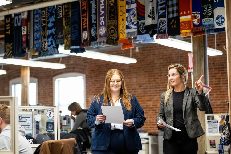 Annmarie Orlando and her mentor Kylie DeWees walk through the Sporting KC office together with rows of soccer scarves hanging from the ceiling rafters above them
